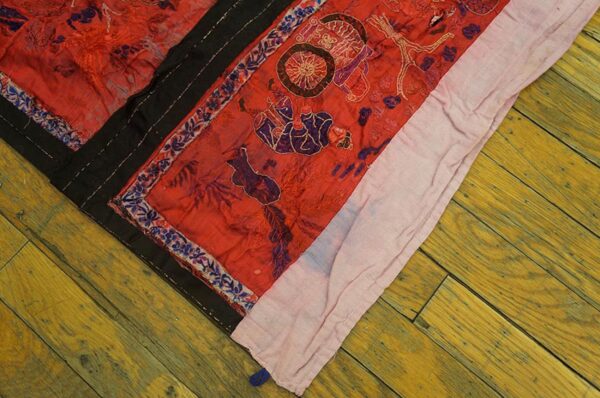 Crinkled red textile with dark purple and gold stitched patterns, black borders, lying on distressed yellow wood floor.