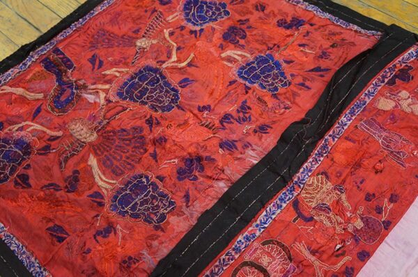 Vibrant red textile featuring elaborate blue floral and gold embroidered bird motifs resting on light wood flooring.