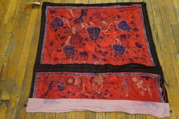 Red embroidered textile with purple florals and black borders lies on distressed wooden flooring.