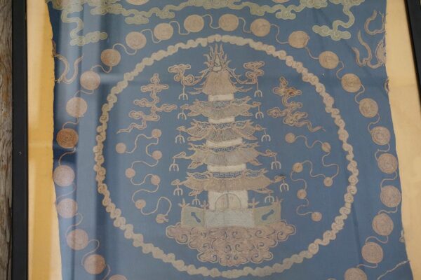 Framed blue textile with embroidered tan pagoda, cloud shapes, and spiraled medallion patterns.