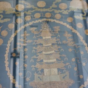 Blue textile with beige embroidered pagoda and cloud motifs, framed and resting on weathered wood.