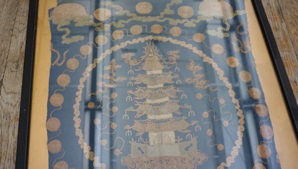 Blue textile with beige embroidered pagoda and cloud motifs, framed and resting on weathered wood.