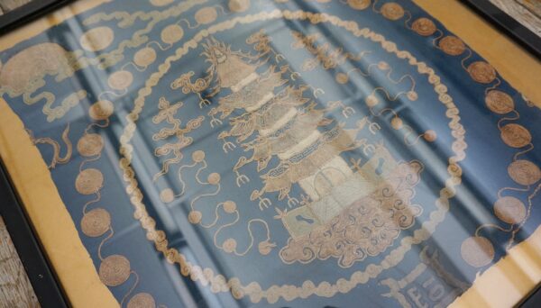 Framed blue textile art features central gold embroidery of a pagoda and surrounding coiled, serpentine patterns.