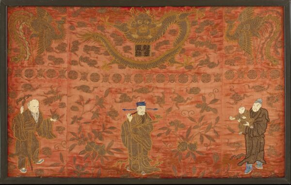 Flat vermilion textile art depicts figures, dragons, and flora embroidered in brown and metallic gold thread within a frame.