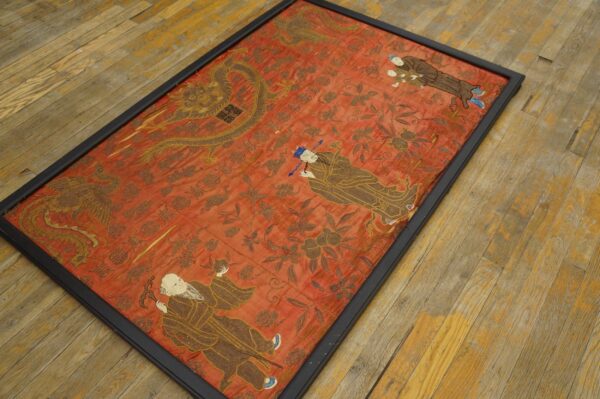 Framed reddish textile featuring gold dragons, phoenix, and figures lies on weathered wood floor.