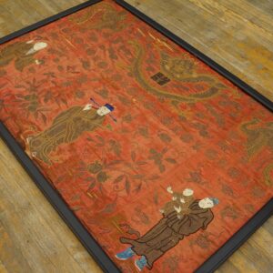 Framed flat red textile featuring detailed brown and gold figures, including a dragon, lying on distressed wood flooring.