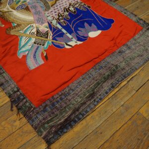 Red embroidered textile with colorful figurative pattern and complex woven borders rests on distressed wood flooring.