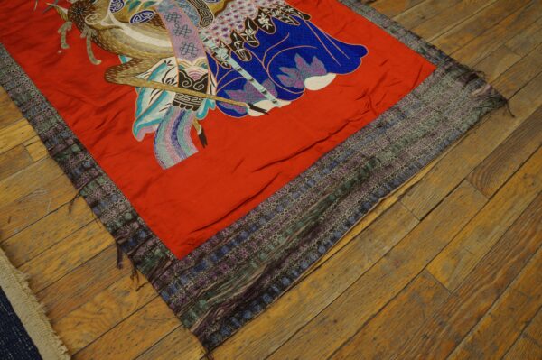 Red embroidered textile with colorful figurative pattern and complex woven borders rests on distressed wood flooring.