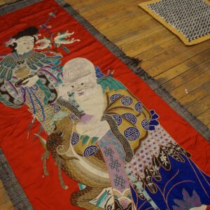 A narrow red embroidered runner featuring detailed figures in teal, purple, and gold rests on a wood floor.