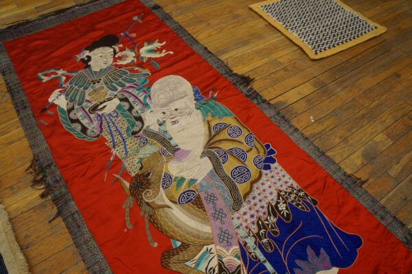 A narrow red embroidered runner featuring detailed figures in teal, purple, and gold rests on a wood floor.