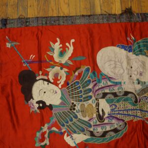 Vibrant red embroidered textile featuring detailed figures and stylized motifs rests on medium brown wood flooring.