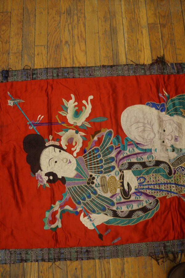 Vibrant red embroidered textile featuring detailed figures and stylized motifs rests on medium brown wood flooring.