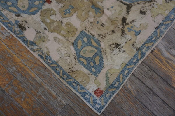 Distressed rug corner with blue border and faded olive designs lies on weathered brown hardwood.