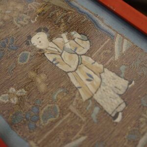 Brown embroidered textile with a cream and blue figure surrounded by stylized floral motifs and a red border.