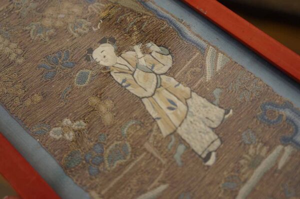 Brown embroidered textile with a cream and blue figure surrounded by stylized floral motifs and a red border.