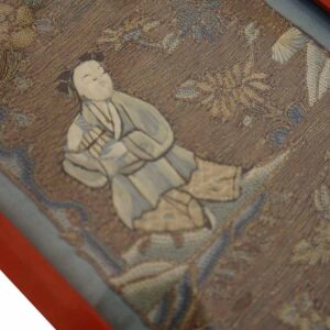 Low-pile textile framed in red shows a beige and blue embroidered figure and dense floral motifs.