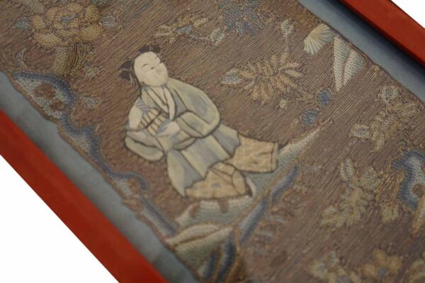 Low-pile textile framed in red shows a beige and blue embroidered figure and dense floral motifs.