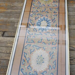 Narrow framed embroidered textile in coral, blue, and green floral patterns rests on weathered wood floors.