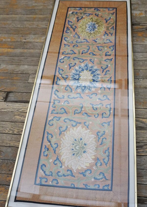 Narrow framed embroidered textile in coral, blue, and green floral patterns rests on weathered wood floors.
