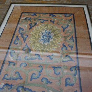 Framed textile features a salmon background with blue and sage floral embroidery, displayed on weathered wooden floorboards.
