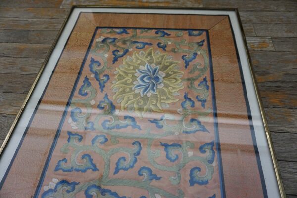 Framed textile features a salmon background with blue and sage floral embroidery, displayed on weathered wooden floorboards.