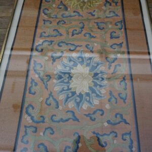 Framed embroidery with blue and green scrolls and a central white flower on a salmon ground.