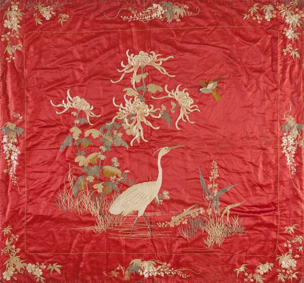 A square, bright red embroidered textile features a white crane, large white flowers, and a small bird within a dense floral border.