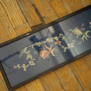 Framed dark blue textile with white and red fish and floral designs resting on distressed yellow-orange wood planks.