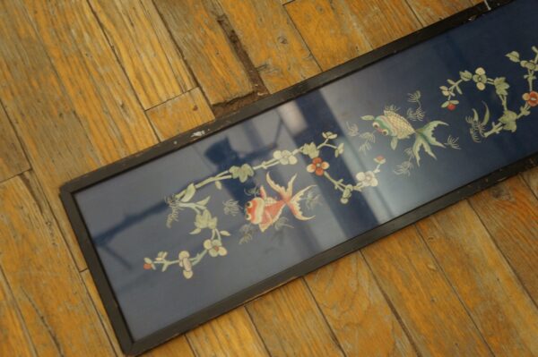 Framed dark blue textile with white and red fish and floral designs resting on distressed yellow-orange wood planks.