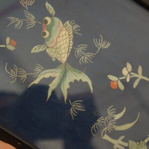 Detailed embroidery features a green and cream fish surrounded by flowers on a dark blue textile.