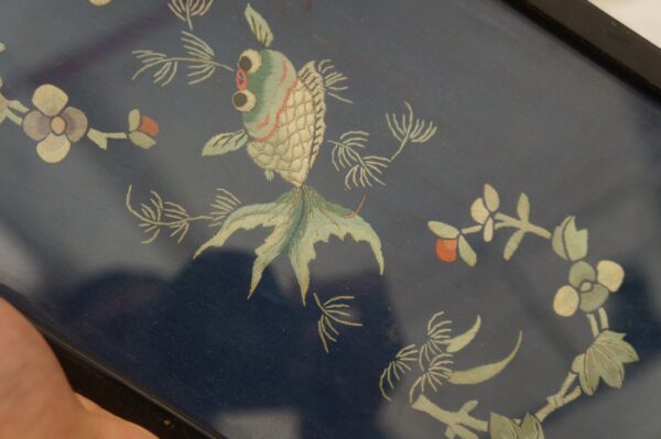 Detailed embroidery features a green and cream fish surrounded by flowers on a dark blue textile.