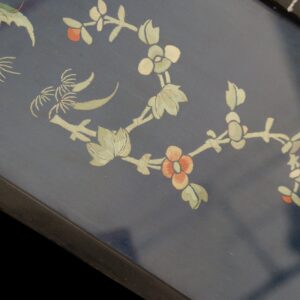 Navy textile with detailed embroidered botanical motifs in pale green and coral, bordered by dark wood.