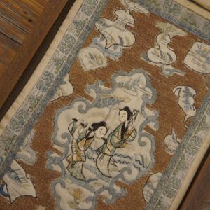 A framed textile featuring brown, cream, and blue embroidery with central figures rests on wood planks.