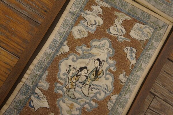 A framed textile featuring brown, cream, and blue embroidery with central figures rests on wood planks.