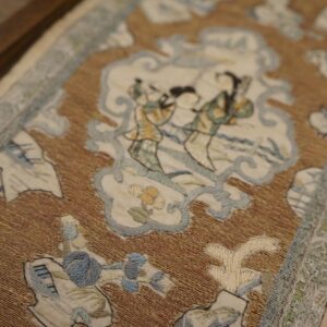 Framed textile featuring a shimmery brown and gold background with elaborate cream, blue, and green figurative scenes.