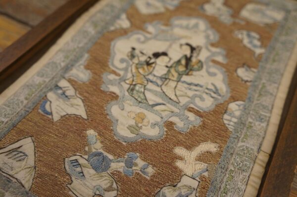 Framed textile featuring a shimmery brown and gold background with elaborate cream, blue, and green figurative scenes.