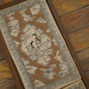 A framed rust and blue flat-weave textile showing two figures, resting on rustic wooden floorboards.
