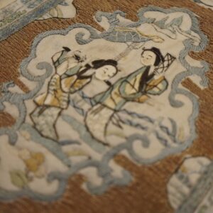 Embroidered scene featuring two figures in cream and soft blue, highlighted by a textured golden-brown background.