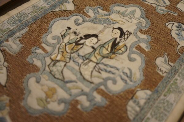 Embroidered scene featuring two figures in cream and soft blue, highlighted by a textured golden-brown background.