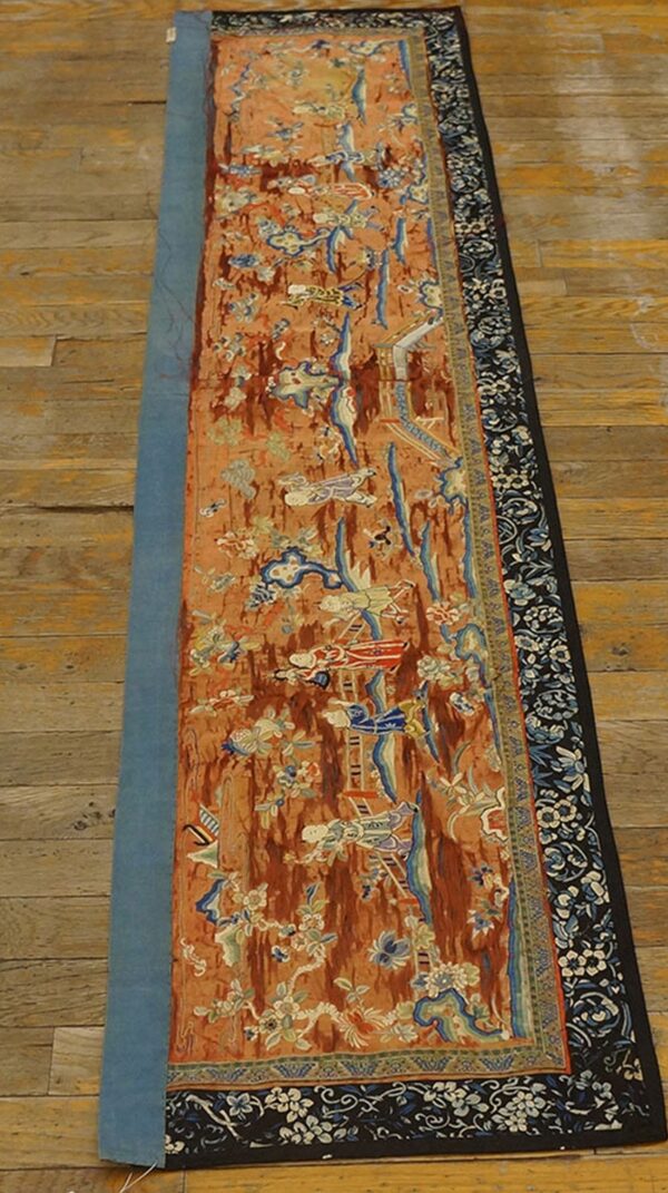 Long, narrow orange and blue textile runner featuring figural patterns, set upon light wood flooring.