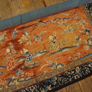 Orange embroidered runner featuring children and blue floral motifs, bordered by dark blue and black, on wood planks.