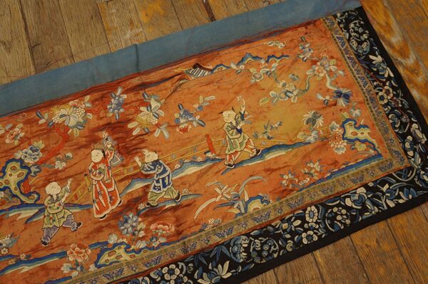 Orange embroidered runner featuring children and blue floral motifs, bordered by dark blue and black, on wood planks.