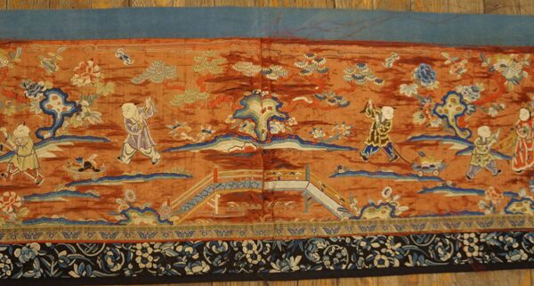 Low-pile tapestry with sienna ground and detailed scene of children, bridge, trees, and a black and blue floral border.