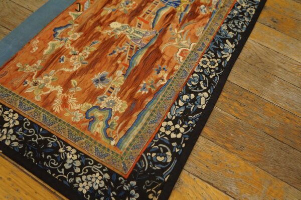 Detailed view of a reddish-orange embroidered textile with blue and white florals resting on wood flooring.