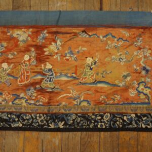 Rust-toned textile panel with blue, white, and green figures and floral patterns, displayed on wood planks.