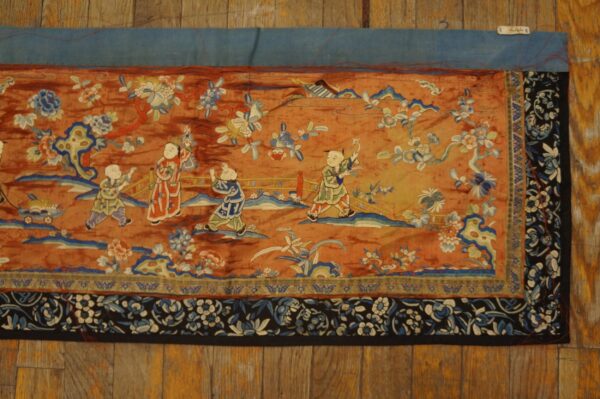 Rust-toned textile panel with blue, white, and green figures and floral patterns, displayed on wood planks.