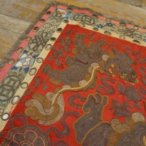 Detailed red textile with coiled brown animal embroidery and mirrored floral borders rests on rustic wood flooring.