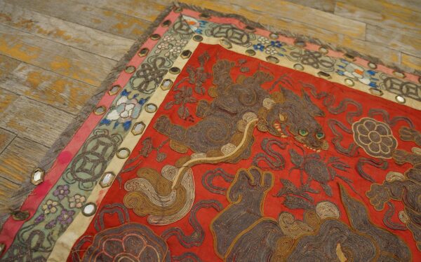 Detailed red textile with coiled brown animal embroidery and mirrored floral borders rests on rustic wood flooring.