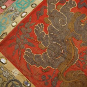 Close-up of a red textile with coiled gold and brown embroidery, segmented by mirrored panels in tan and teal.