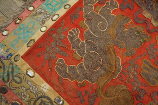 Close-up of a red textile with coiled gold and brown embroidery, segmented by mirrored panels in tan and teal.